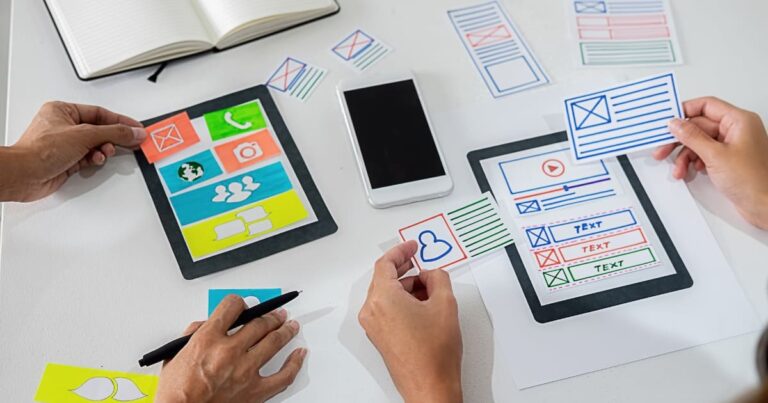 Personas diseñando la estructura visual de una aplicación móvil con elementos gráficos y plantillas impresas.