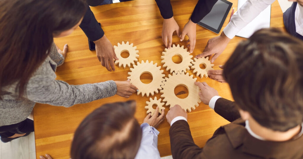 Personas colaborando alrededor de engranajes de madera como símbolo de trabajo en equipo e innovación sin presupuesto.